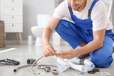 Plombier professionnel en vêtements de travail (jean et polo de couleur neutre avec logo discret) en train de réparer une robinetterie ou une chasse d'eau dans une salle de bain résidentielle. Le plombier est photographié de côté, concentré sur son travail, outillage adapté à la main (clé, tournevis). Arrière-plan flou montrant une salle de bain classique. Lumière naturelle, atmosphère professionnelle et rassurante, sans pose artificielle.