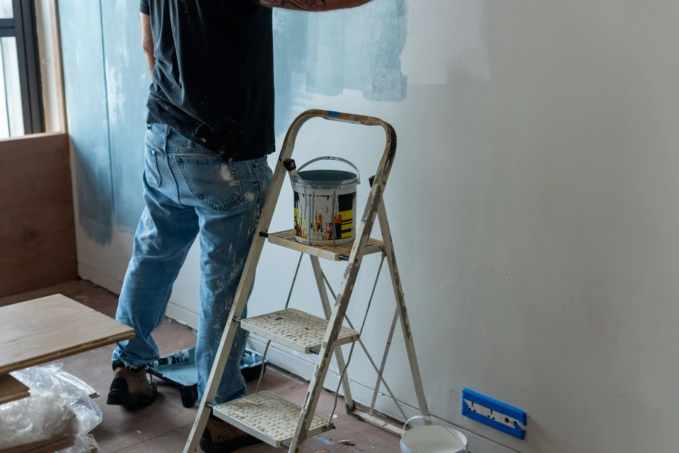 Photographie d'un chantier intérieur de rénovation de salle de bains, montrant plusieurs artisans ou intervenants travaillant simultanément en coordination : un carreleur en train de poser du carrelage, un électricien installer une colonne thermostatique de douche (ou des conduits électriques), un peintre ou autre intervenant en arrière-plan, tous opérant dans le même espace, symbolisant la coordination intégrée de multiples corps de métier sous la supervision d'une même équipe. La scène doit être organisée et efficace, sans confusion.